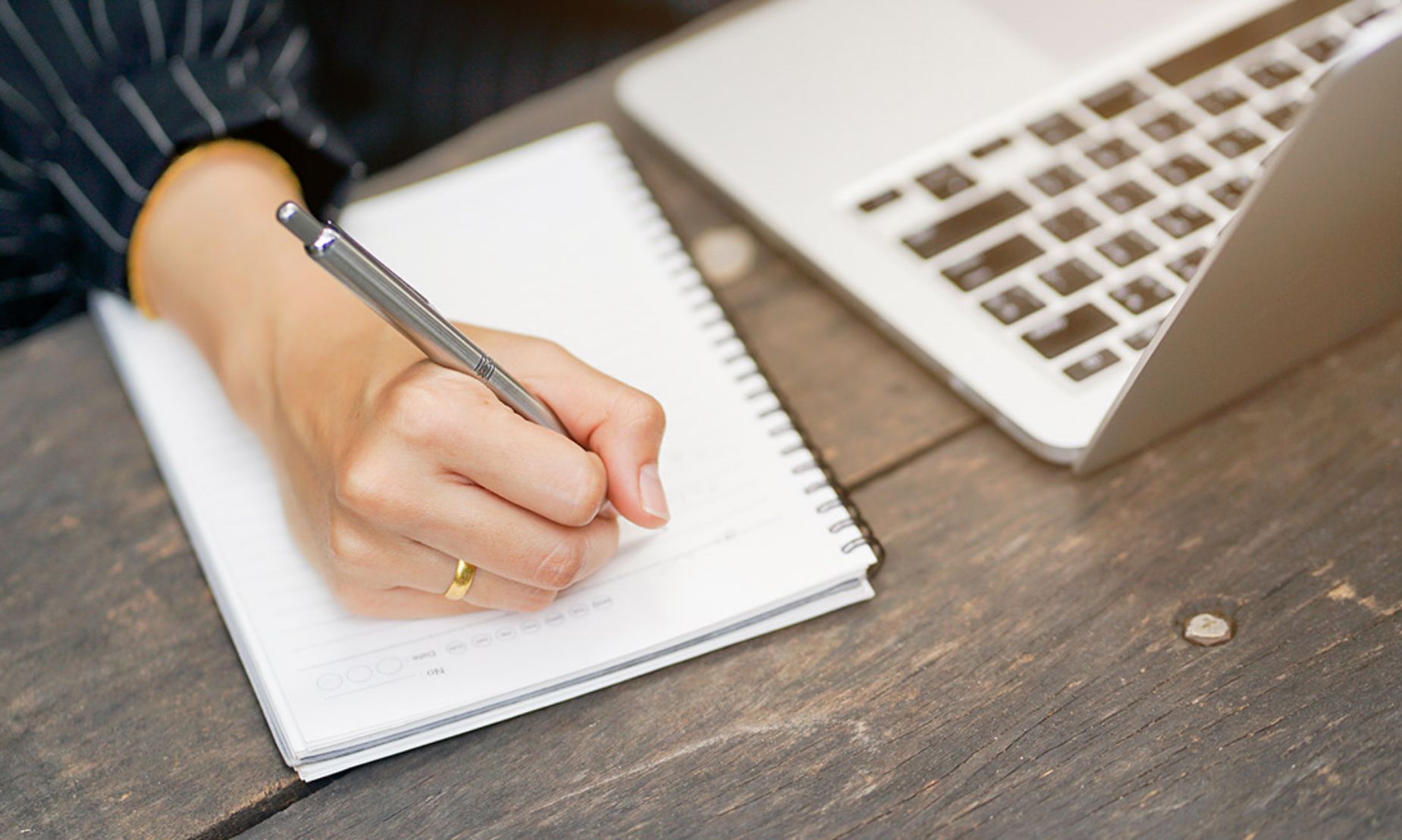 Close-up of a hand writing in a spiral notebook beside an open laptop on a wooden desk, warm neutral calm focus